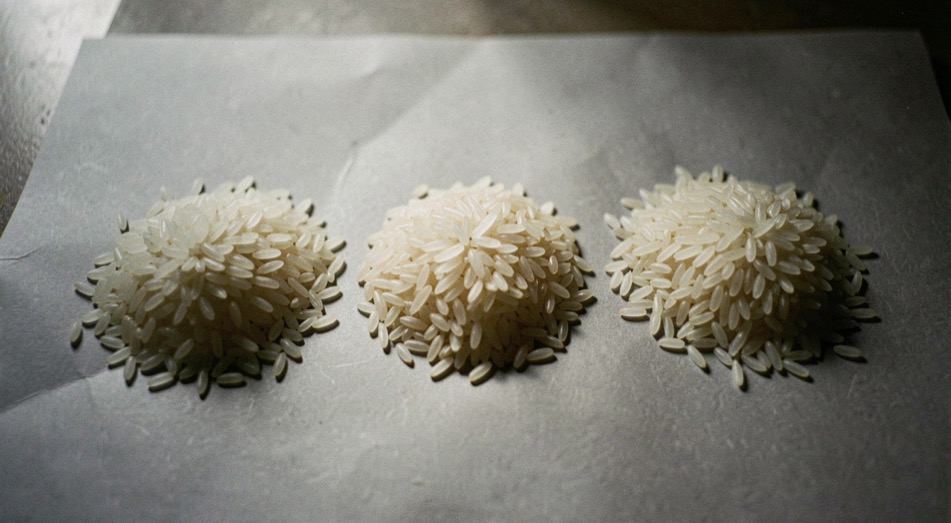 Trois petits tas de grains de riz blanc de tailles et de formes différentes alignés sur une feuille de papier gris, éclairage rasant qui souligne le relief des grains